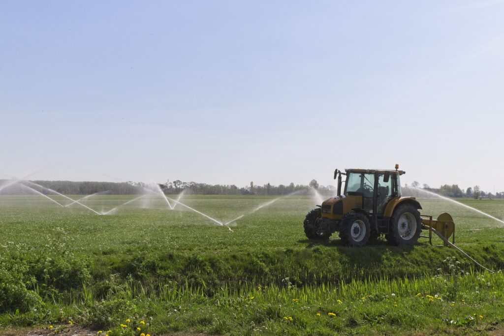 Bilden visar en traktor och vattenspridare på en åker. Hitta traktordelar och traktortillbehör som harv, slåtter och snökedjor till traktorer som exempelvis till Massey Ferguson.